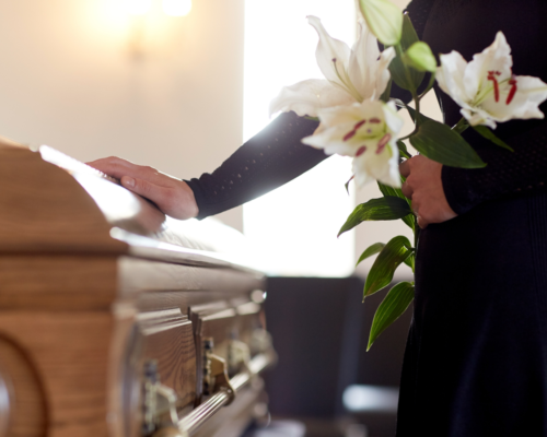 Man in black placing his hand on a casket and holding flowers in another hand at a funeral.